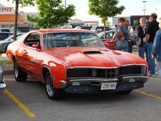 an orange muscle car parked in a parking lot with people standing around and looking at it