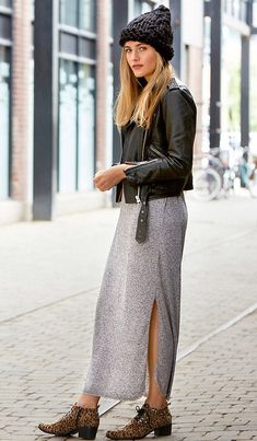 a woman standing on the sidewalk wearing a black hat and grey skirt with side slit