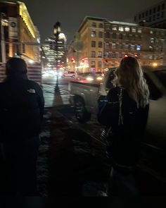 two people standing in the street at night with buildings and lights behind them, one person is on her cell phone