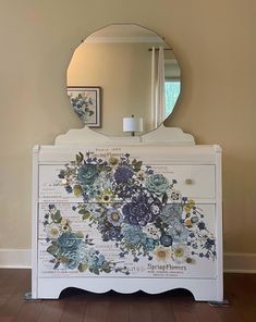 a white dresser with flowers painted on it and a round mirror over the top of it