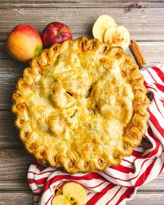 an apple pie on a red and white striped towel next to two apples, cinnamon sticks, and some other fruit