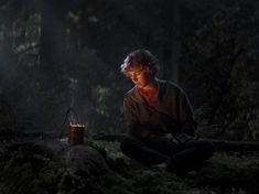 a woman sitting on the ground next to a lantern in the dark forest at night