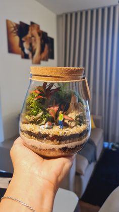 a person holding up a fish bowl filled with plants and small figurines in it