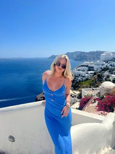 a woman in a blue dress standing on a balcony overlooking the ocean and houses with white walls