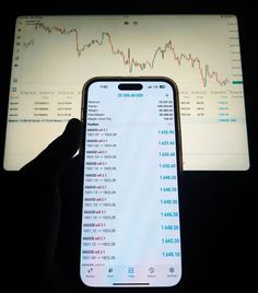 a person holding up an iphone in front of a computer screen with stock prices on it