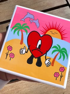 a hand holding up a square painting with a heart on it and palm trees in the background