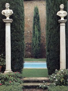 two white pillars with statues on them in front of a pool surrounded by greenery