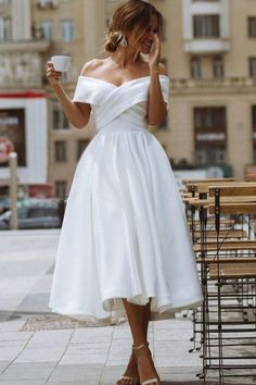 a woman in a white dress holding a cup and talking on her cell phone while walking down the street