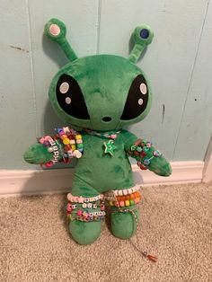 a green stuffed animal with lots of beads on it's legs and eyes sitting against a wall