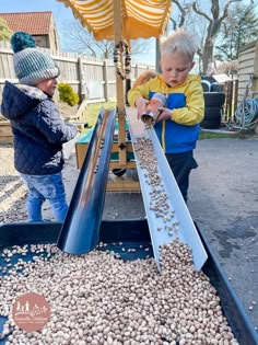 two young boys are playing with peanuts in an outdoor play area that is filled with sand and gravel