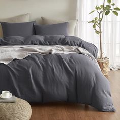 a bed with grey linens and pillows in a white room next to a potted plant