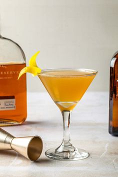 an orange cocktail garnished with a lemon peel in a martini glass next to two bottles