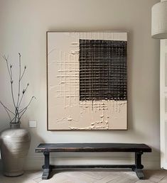 an abstract painting hangs on the wall next to a bench and vase with flowers in it