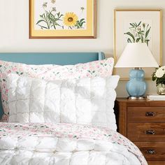 a bed with white comforter and pillows in a bedroom next to a lamp on a dresser