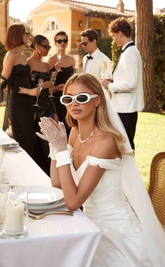 a woman in white dress and sunglasses sitting at a table with other people around her