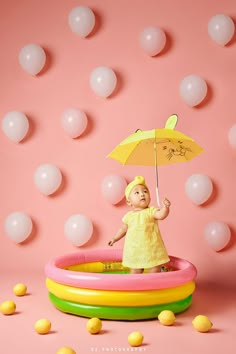 a baby girl holding an umbrella in a pool with balloons floating around her on a pink background