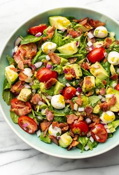 a salad with bacon, avocado and tomatoes in a bowl on a marble table