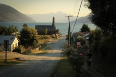 a person walking down a dirt road next to the ocean and houses with mountains in the background