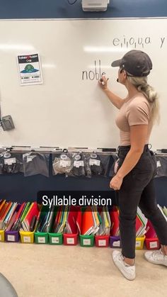 a woman writing on a whiteboard in front of a classroom full of school supplies