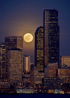 the full moon is seen in the sky over some tall buildings and skyscrapers at night