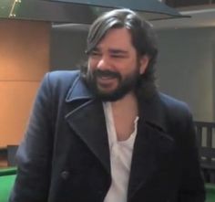 a man with long hair and beard standing in front of a pool table smiling at the camera