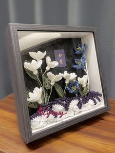 a frame with crocheted flowers in it sitting on a wooden table next to a wall