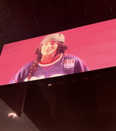 a woman with long hair wearing a baseball cap and standing in front of a large screen