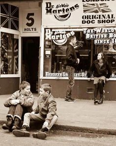 black and white photograph of two boys sitting on the sidewalk in front of a store