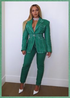 a woman wearing a green suit and white shoes standing in front of a wall with her hands on her hips