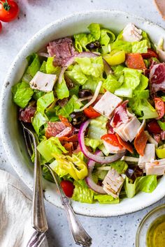 a white bowl filled with salad next to two spoons and tomatoes on the side