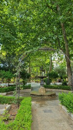 a park with trees, benches and a fountain in the foreground is surrounded by greenery