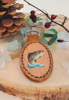 a leather keychain with a fish on it sitting next to a pine cone