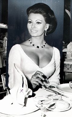 a black and white photo of a woman sitting at a table with wine glasses in front of her