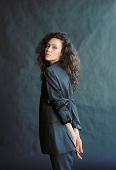 a woman with curly hair standing in front of a dark background wearing a black suit