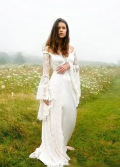 a woman standing in a field wearing a white dress with long sleeves and an open shoulder