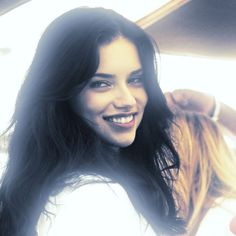 a woman with long black hair smiling and holding her hand up to her head while sitting in a car