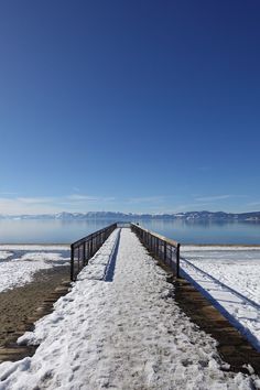 a snow covered walkway leading to the water on a sunny day with blue skies and mountains in the background