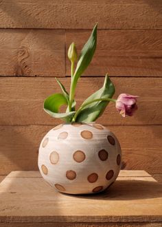 a white vase with brown spots and a pink flower in it on a wooden table