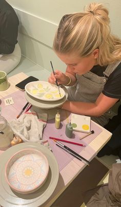 a woman is painting on a plate with paintbrushes