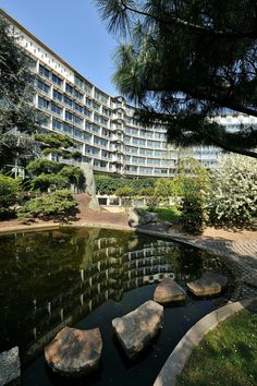 a pond in front of a large building with trees and bushes around it, surrounded by rocks