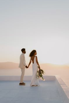 a bride and groom hold hands as the sun sets in the distance behind them on top of a building