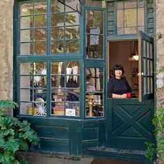 a woman standing in the doorway of a store with green shutters and glass windows