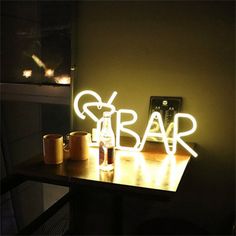 a wooden table topped with two cups next to a neon sign that says bar on it