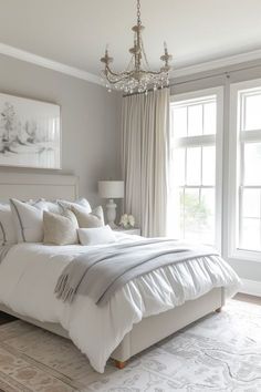 a large white bed sitting in a bedroom next to two windows and a chandelier