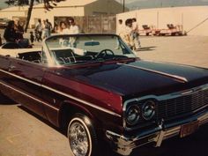 an old car parked in a parking lot with people walking around it and onlookers