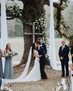 a bride and groom are kissing under the chute at their outdoor wedding ceremony in front of an oak tree