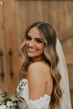 a woman in a wedding dress holding a bouquet and smiling at the camera while standing next to a wooden wall