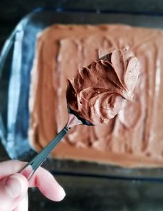 a person holding a spoon with chocolate frosting on it in front of a square cake