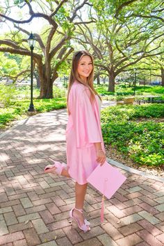 a woman in pink holding a pink purse and standing on brick walkway with trees behind her