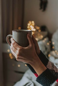 a person holding a coffee cup in their hand while sitting on a bed next to a christmas tree